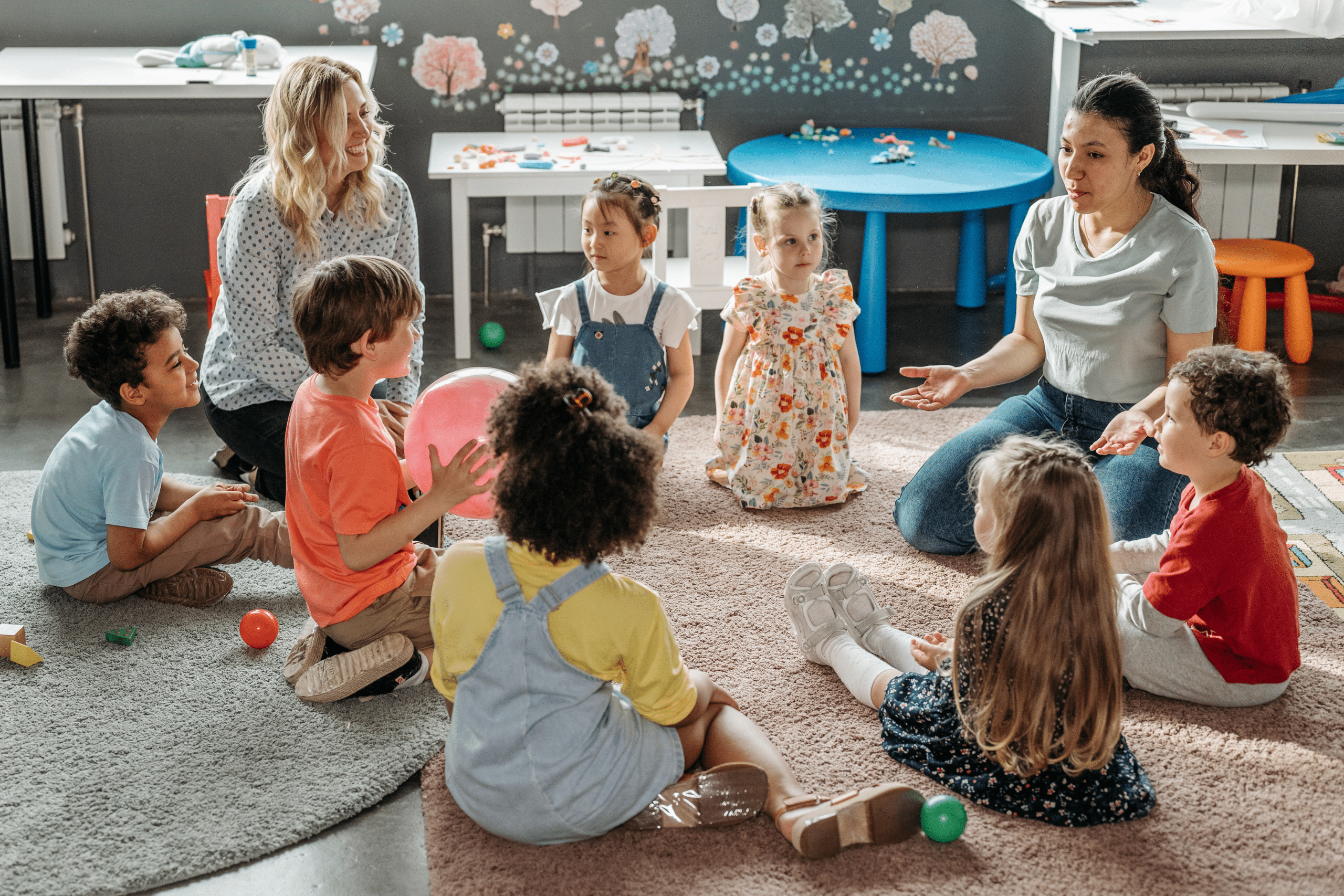 A photo of teachers and students sitting in a circle on a rug, passing a ball back and forth.